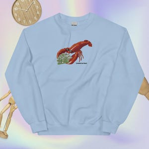 May include: Light blue sweatshirt featuring a red lobster holding a wad of cash. The text "Come get tax fraud." is printed below the lobster.
