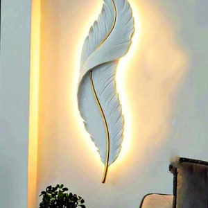 Könnte beinhalten: Weiße wandleuchte in federform mit goldener akzentuierung, beleuchtet mit warmweißem licht. Die wandleuchte ist an einer weißen wand montiert.