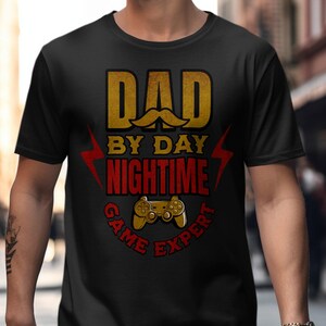 May include: Black t-shirt with a graphic design that says "Dad by Day Nighttime Game Expert" with a gold and red color scheme and a stylized video game controller.