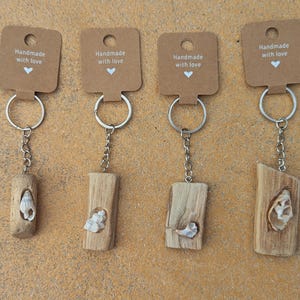 Könnte beinhalten: Eine Sammlung von sechs Schlüsselanhängern mit Anhängern aus Naturholz, jeweils mit einem einzigartigen Schnitzdesign und einer kleinen Muschel. Die Schlüsselanhänger haben silberfarbene Ringe und Ketten und sind an braunen Anhängern mit der Aufschrift "Handmade with love" befestigt.