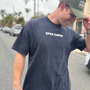 May include: A person wearing a dark blue t-shirt with the text "SPKR HMPRZ" printed in white. The person is also wearing a green mesh baseball cap with the text "ORAC Red Bull RAC" printed on it.
