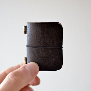 Op de afbeelding: Een klein, donkerbruin leren notitieboekje met een getextureerd oppervlak. Het notitieboekje wordt bij elkaar gehouden door een zwarte elastische band en heeft metalen hoekaccenten. Het notitieboekje is compact en lijkt een reisdagboek te zijn.