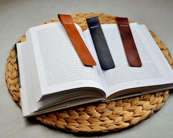 May include: Three leather bookmarks in brown, dark brown, and blue, lying on a book open on a woven straw mat.