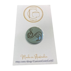 May include: A round, silver-coloured coin with a brown treble clef and the letter "D" engraved on it. The coin is on a white card with the text "Custom Coin Co." and "Made in Australia" printed on it.