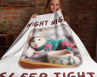 Puede incluir: Una manta de forro polar blanca con un borde marrón y el texto "Night Night Sleep Tight" impreso en ella. La manta presenta una rata blanca con un suéter a rayas verdes y rojas, acostada en una cama con una taza de chocolate caliente.