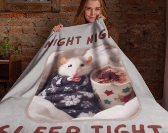 Puede incluir: Una rata blanca con un suéter blanco y negro está acurrucada en una manta con una taza de chocolate caliente. La manta tiene el texto "Night Night Sleep Tight" impreso.