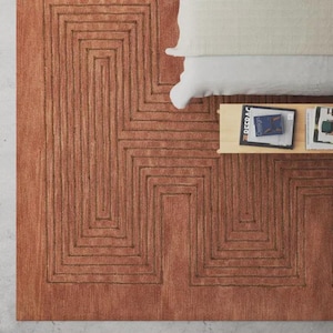 May include: A terracotta-colored rug with a geometric pattern of interlocking rectangles. The rug is placed next to a bed with a white pillow and a small wooden side table with books.