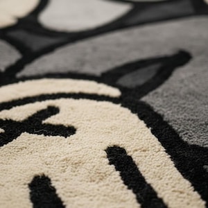 K&ouml;nnte beinhalten: Nahaufnahme eines Teppichs mit einem sich wiederholenden Muster aus stilisierten Cartoon-Charakterelementen. Der Teppich hat eine strukturierte, weiche Oberfl&auml;che mit schwarzen, cremefarbenen und grauen Farben. Das Design umfasst abgerundete Formen und X-f&ouml;rmige Details.