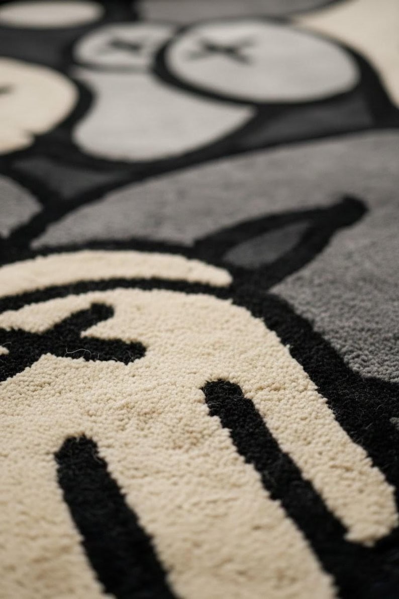 K&ouml;nnte beinhalten: Nahaufnahme eines Teppichs mit einem sich wiederholenden Muster aus stilisierten Cartoon-Charakterelementen. Der Teppich hat eine strukturierte, weiche Oberfl&auml;che mit schwarzen, cremefarbenen und grauen Farben. Das Design umfasst abgerundete Formen und X-f&ouml;rmige Details.