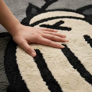 K&ouml;nnte beinhalten: Nahaufnahme eines grau-, schwarz- und cremefarbenen Teppichs mit einem cartoonartigen Design. Der Teppich weist ein sich wiederholendes Muster aus stilisierten Gesichtern und Formen auf. Eine Hand liegt auf dem Teppich und zeigt die Textur des Materials.