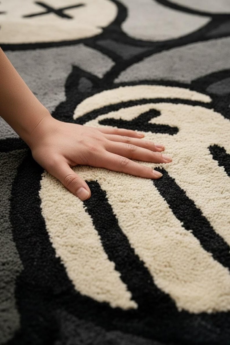 K&ouml;nnte beinhalten: Nahaufnahme eines grau-, schwarz- und cremefarbenen Teppichs mit einem cartoonartigen Design. Der Teppich weist ein sich wiederholendes Muster aus stilisierten Gesichtern und Formen auf. Eine Hand liegt auf dem Teppich und zeigt die Textur des Materials.