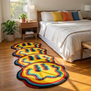 May include: Three colourful, trippy smiley face rugs on a hardwood floor. The rugs have a yellow base with red, blue, and black borders. The rugs are in front of a bed with white bedding.