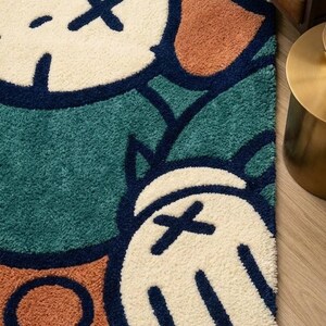 K&ouml;nnte beinhalten: Nahaufnahme eines farbenfrohen Teppichs mit einem Cartoon-Charakter-Design. Der Teppich hat einen t&uuml;rkisgr&uuml;nen Hintergrund mit cremefarbenen Formen und orangefarbenen und marineblauen Akzenten. Der Teppich besteht aus einem weichen, strukturierten Material.