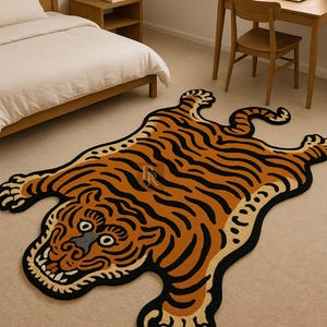 Op de afbeelding: Een decoratief tapijt in de vorm van een tijger, met oranje en zwarte strepen op een beige achtergrond. Het tapijt heeft een zwarte rand en ligt op een beige tapijt. De tijgerkop is gedetailleerd met ogen, een neus en een mond. Ongeveer 180 cm lang.