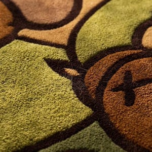 Pode incluir: Close-up de um tapete texturizado com um padr&atilde;o repetitivo em tons de castanho e verde. O design apresenta formas arredondadas com contornos castanhos escuros e um detalhe em forma de cruz.