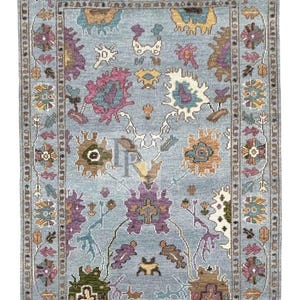 K&ouml;nnte beinhalten: Ein blauer Teppich mit einem floralen Muster in den Farben Rosa, Lila, Braun und Gr&uuml;n. Der Teppich hat einen Fransenrand.