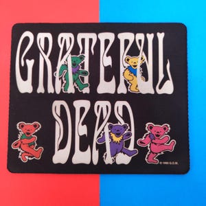 May include: Black rectangular coaster with the words "Grateful Dead" in white, featuring colourful dancing bears. The bears are in shades of red, green, yellow, purple, and pink. The coaster has a red and blue background.