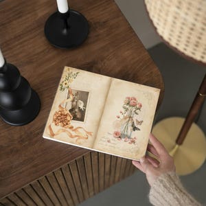 K&ouml;nnte beinhalten: Ein offenes Journal mit Illustrationen im Vintage-Stil von Blumen, Schmetterlingen und einer Geige. Das Journal wird von einer Person gehalten und liegt auf einer dunklen Holzoberfl&auml;che. Ein schwarzer Kerzenhalter mit einer wei&szlig;en Kerze ist im Hintergrund zu sehen.