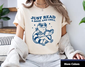 Może przedstawiać: Kremowa koszulka unisex z krótkim rękawem z dżerseju z niebieskim napisem "JUST READ A BOOK AND CHILL". Koszulka przedstawia niebieską grafikę żaby w okularach przeciwsłonecznych i bikini, czytającej książkę na kole ratunkowym.