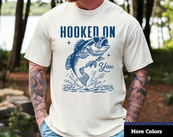 May include: A cream-colored unisex t-shirt with a blue graphic of a leaping fish with the words "HOOKED ON YOU". The shirt also has the text "1-2-TEE CREATIVE" and "Comfort Colors 1717".