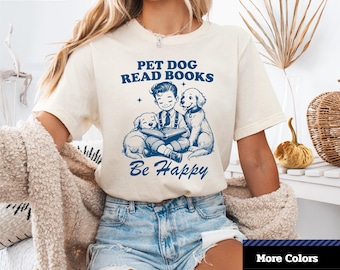 Può includere: T-shirt unisex color crema a maniche corte con grafica blu. La grafica raffigura un ragazzo che legge a due cani, con la scritta "PET DOG READ BOOKS Be Happy". La maglietta ha il logo "1-2-TEE CREATIVE" in alto a sinistra.