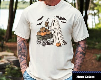May include: Cream-colored unisex dyed t-shirt with a Halloween-themed graphic. The design features a ghost pushing a cart filled with pumpkins, holding a coffee, and surrounded by bats. The shirt has the text "Comfort Colors 1717".