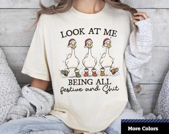 May include: Cream-colored unisex dyed t-shirt with a humorous graphic. The graphic features three cartoon geese wearing Santa hats and boots, with the text "LOOK AT ME BEING ALL festive and Shit."