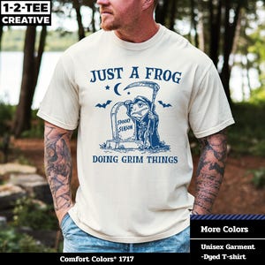 Könnte beinhalten: Cremefarbenes Unisex-T-Shirt mit einem blauen Aufdruck, der einen Frosch als Sensenmann zeigt, der neben einem Grabstein mit der Aufschrift "Spooky Season" steht. Der Text "Just a Frog Doing Grim Things" ist ebenfalls auf dem Shirt aufgedruckt.