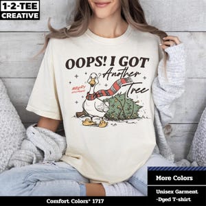 Puede incluir: Camiseta unisex color crema con un diseño de un ganso con bufanda y un árbol de Navidad. El texto dice "OOPS! I GOT Another Tree" y "MERRY GOOSEMAS". La camiseta es una camiseta teñida Comfort Colors. El logotipo de la marca también es visible.
