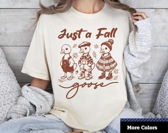 May include: Cream-colored unisex dyed t-shirt with a brown graphic of three ducks dressed in fall attire, with the text "Just a Fall goose". The shirt has the text "Comfort Colors 1717" and "More Colors" on the bottom.