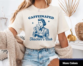 Könnte beinhalten: Cremefarbenes Unisex-Jersey-Kurzarm-T-Shirt mit dem Text "CAFFEINATED Teacher's Club" und einer blauen Illustration einer Frau, die eine Kaffeetasse hält. Das Shirt hat einen Rundhalsausschnitt und ist aus weichem Material.