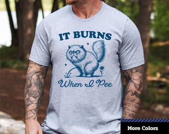 May include: A light gray unisex jersey short sleeve tee with the text "IT BURNS When I Pee" above and below a cartoon cat urinating. The shirt also has the logo "1-2-TEE CREATIVE" in the upper left corner.