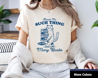 May include: Cream-colored unisex jersey short sleeve tee with the text "There's No Such Thing As Too Many Books" and a blue illustration of a cat carrying books. The shirt also has the "1-2-TEE CREATIVE" logo.