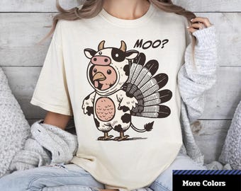 Puede incluir: Camiseta unisex teñida de color crema con un personaje de dibujos animados híbrido vaca-pavo. El personaje tiene una cabeza de vaca y un cuerpo de pavo, con la palabra "MOO?" encima. La camiseta tiene el texto "Comfort Colors 1717" y "More Colors".