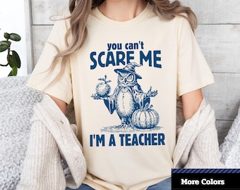 May include: Cream-colored unisex short sleeve tee with a blue graphic of an owl wearing a witch hat, holding an apple and a pumpkin. Text reads "you can't scare me, I'm a teacher." The shirt has the "1-2-TEE CREATIVE" logo in the top left corner.
