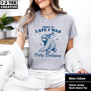 May include: A light gray unisex jersey short sleeve tee featuring a blue illustration of a sloth wearing a party hat. The text on the shirt reads "Sorry I'm Late I Was Dilly Dallying."