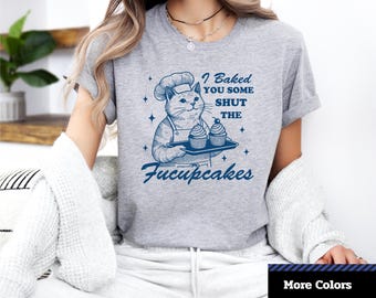Puede incluir: Camiseta unisex de manga corta de color gris claro con un gato chef sosteniendo cupcakes. La camiseta tiene el texto "I Baked You Some Shut The Fucupcakes" en azul. También se ve el logo "1-2-TEE CREATIVE".