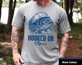 May include: A gray unisex dyed t-shirt with a blue graphic of a fish wearing a helmet and smoking a pipe. The text "HOOKED ON YOU" is below the graphic. The shirt is a Comfort Colors 1717.