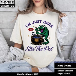 Puede incluir: Camiseta unisex color crema con un gráfico de una rana verde removiendo en una olla. El texto dice "I'M JUST HERE TO Stir The Pot". La camiseta también tiene el texto "Comfort Colors 1717", "More Colors" y "Unisex Garment -Dyed T-shirt".