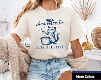 Op de afbeelding: Crèmekleurig unisex T-shirt met korte mouwen met de tekst "I'M Just Here To STIR THE POT" en een blauwe illustratie van een kat die in een pot roert. Het shirt heeft ook het logo "1-2-TEE CREATIVE".