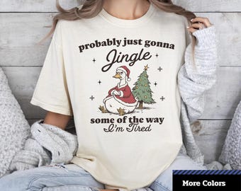 May include: Cream-colored unisex t-shirt with a Christmas-themed graphic. The graphic features a duck wearing a Santa hat and suit, with the text "probably just gonna Jingle some of the way I'm Tired". The shirt also has the text "Comfort Colors 1717".