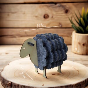 Könnte beinhalten: Eine Schaf-Figur aus Holz und Filz. Das Schaf hat einen hellgrünen Kopf und Beine mit einem blauen Filzkörper. Die Figur ist 14,5 cm lang, 11 cm hoch und 10 cm breit. Das Schaf wird auf einer Holzscheibe präsentiert.
