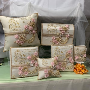Op de afbeelding: Een verzameling ivoorkleurige decoratieve items, waaronder kussens en dozen, versierd met bloemstukken, vlinders en gouden accenten. De tekst "Mis Quince Años" is in gouden letters weergegeven, ter viering van een quinceañera.