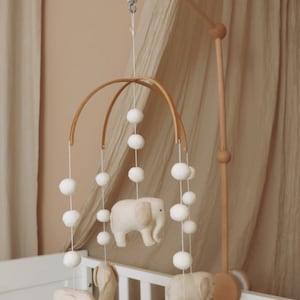 May include: A wooden baby mobile with a white musical box and five beige felt elephant toys hanging from it. The mobile is suspended from a wooden arm and is attached to a white crib.