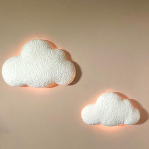Peut inclure: Deux appliques murales en forme de nuage, blanches et duveteuses. Elles émettent une lumière chaude et douce, créant une ambiance chaleureuse. Fixées sur un mur rose pâle, elles apportent une touche de fantaisie à la décoration intérieure. Idéales pour une chambre d'enfant.