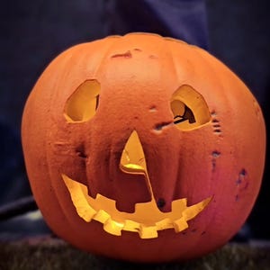 Könnte beinhalten: Ein geschnitzter orangefarbener Kürbis mit einem leuchtenden Jack-o'-Lantern-Gesicht, dreieckigen Augen und einem gezackten Mund. Unter dem Kürbis zeigt ein teilweises Schild die Buchstaben WEEN und den Namen @cellar_toys in weißer Schrift.