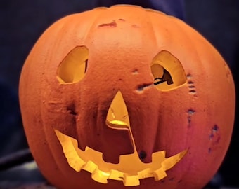 May include: A carved orange pumpkin with a glowing jack-o'-lantern face featuring triangular eyes and a jagged mouth. Below the pumpkin, a partial sign displays the letters WEEN and the social media handle @cellar_toys in white text.