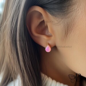 May include: A gold-colored earring with a teardrop-shaped pink opal stone. The earring is set in a delicate gold frame with a beaded edge. The earring is shown on a person's ear.