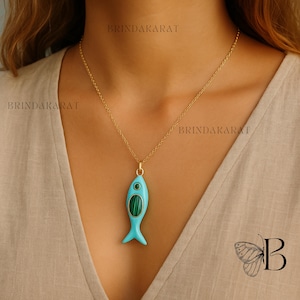 May include: A gold chain necklace with a turquoise fish pendant. The fish pendant has a malachite inlay and a small green stone eye. The necklace is worn against a neutral-toned top.