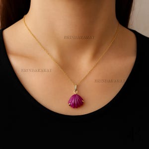 Può includere: Una collana color oro con un ciondolo a forma di conchiglia magenta. Il ciondolo è appeso a una delicata catena. La collana è indossata su un top nero. Il testo "BRINDAKARAT" è visibile sullo sfondo.